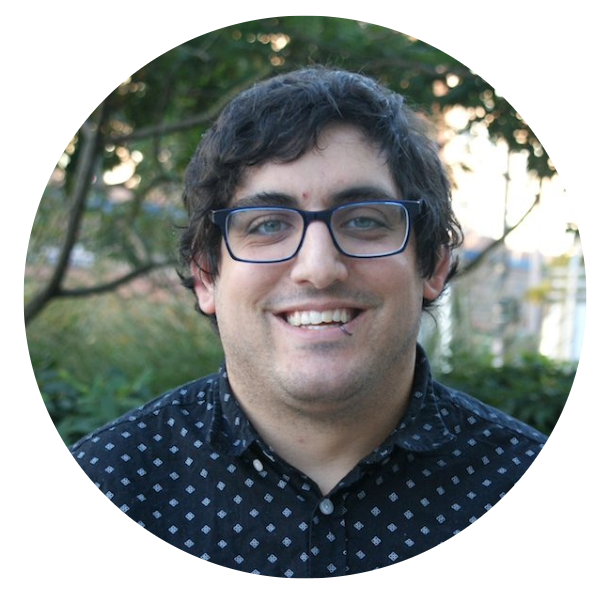 A man with curly dark hair wearing glasses and dotted shirt smiles at camera.