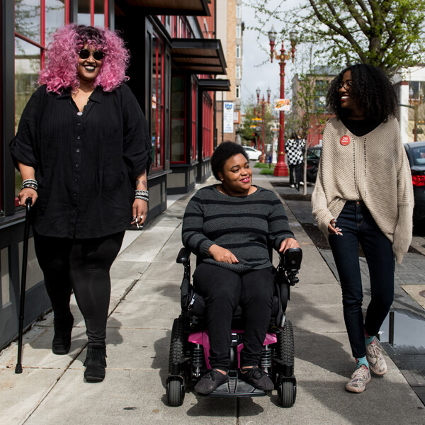 Three people on sidewalk. Person on the left is using a cane. Person in middle is in a wheelchair.