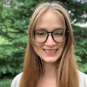 Photo of Charla wearing black rimmed glasses, with light brown hair parted in the middle. She is wearing a white top and standing in front of green bushes.
