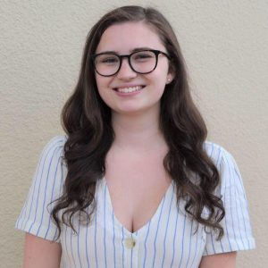 Photo of Lily wearing a white striped short sleeve shirt. She is wearing black rimmed glasses and has long brown curled hair.