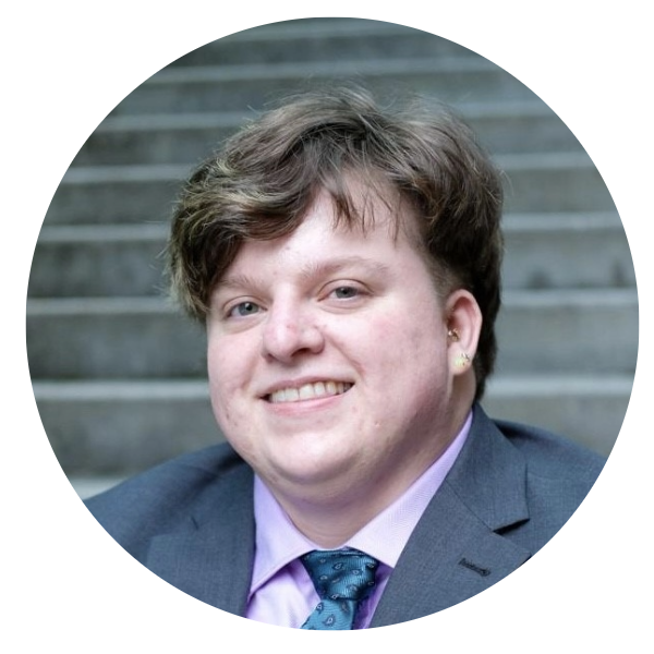 A white person with short dirty blonde hair wearing a grey blazer and blue tie.