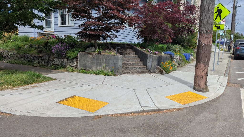The focus of the photo is a street corner with two curb ramps. In the background there is a colorful garden with concrete stairs leading up to a blue house